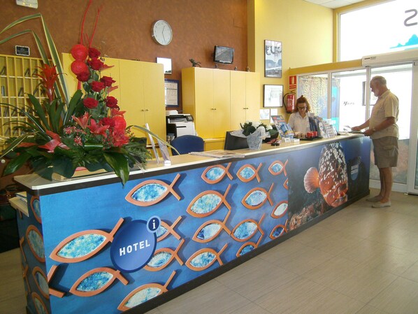 A hotel reception desk with a large fish-themed mural, a clock, and a fire extinguisher on the wall.
