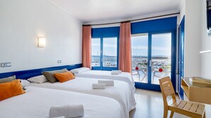 Habitación de hotel con dos camas, un escritorio y una silla. Amplia ventana con vista a un puerto deportivo y edificios.