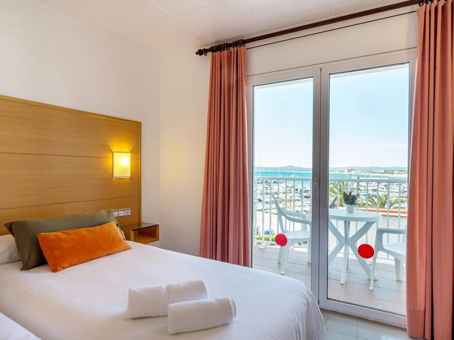 Habitación de hotel con cama, mesita de noche, lámpara, balcón con mesa y sillas, y vista al mar y edificios.