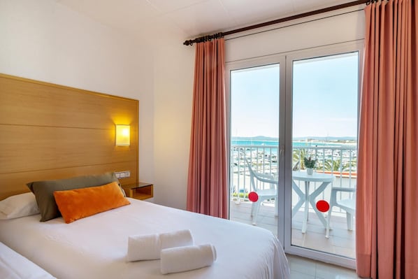 Habitación de hotel con cama, mesita de noche, lámpara, balcón con mesa y sillas, y vista al mar y edificios.
