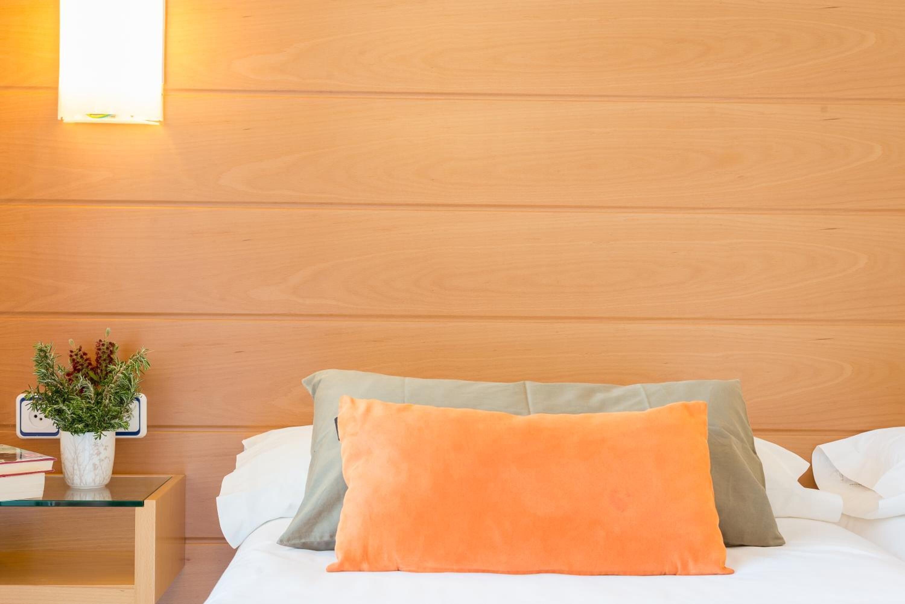 A neatly made bed with two pillows, a bedside table with a plant, and a wall-mounted light.