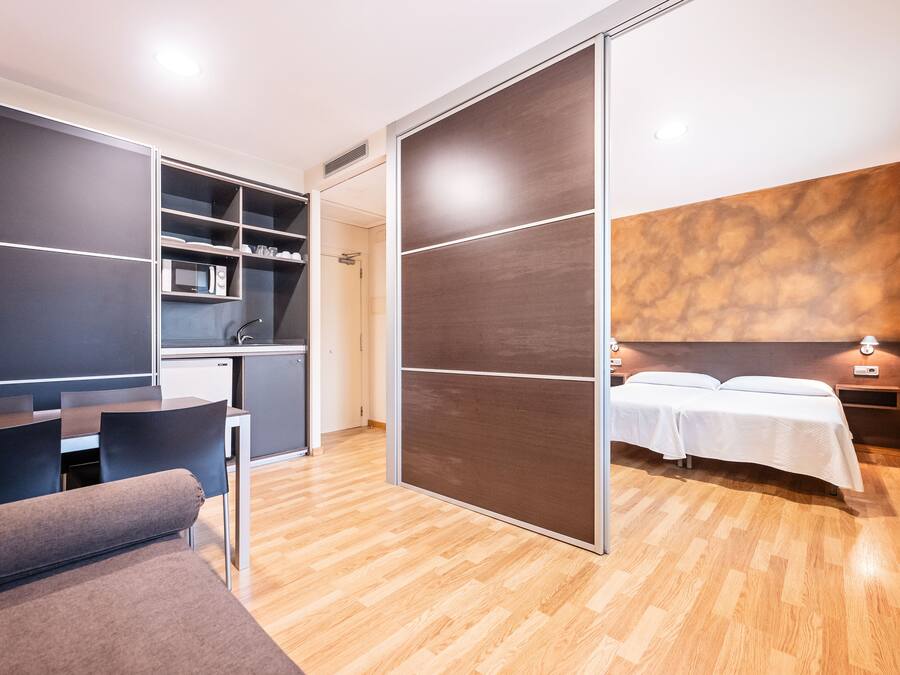Una habitación de hotel moderna con suelo de madera, un sofá, una cama con ropa blanca, un escritorio y una kitchenette.