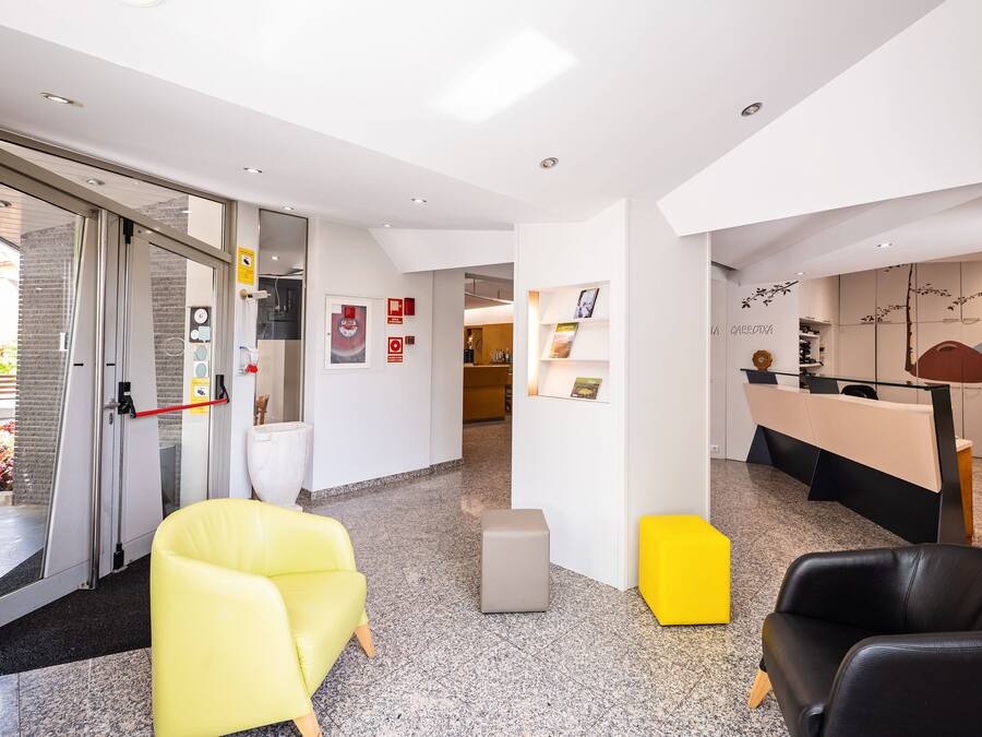 Un espace d’accueil moderne doté d’une porte d’entrée en verre, d’un comptoir de réception et de différents sièges, notamment un fauteuil jaune et un fauteuil en cuir noir.