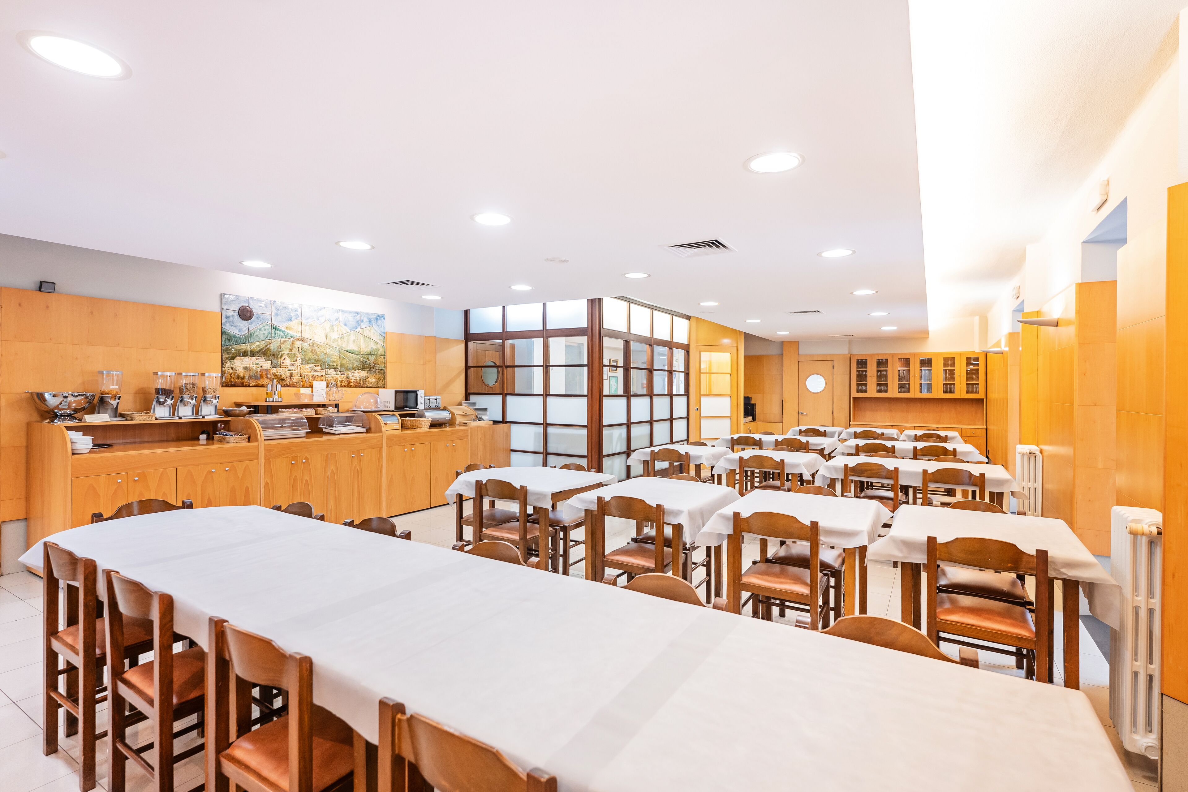 Un comedor amplio con varias mesas y sillas, una estación de buffet y un gran ventanal que proporciona luz natural.