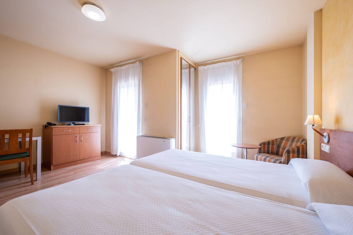 Habitación de hotel con dos camas, un televisor sobre un mueble de madera, una mesita pequeña y una silla.