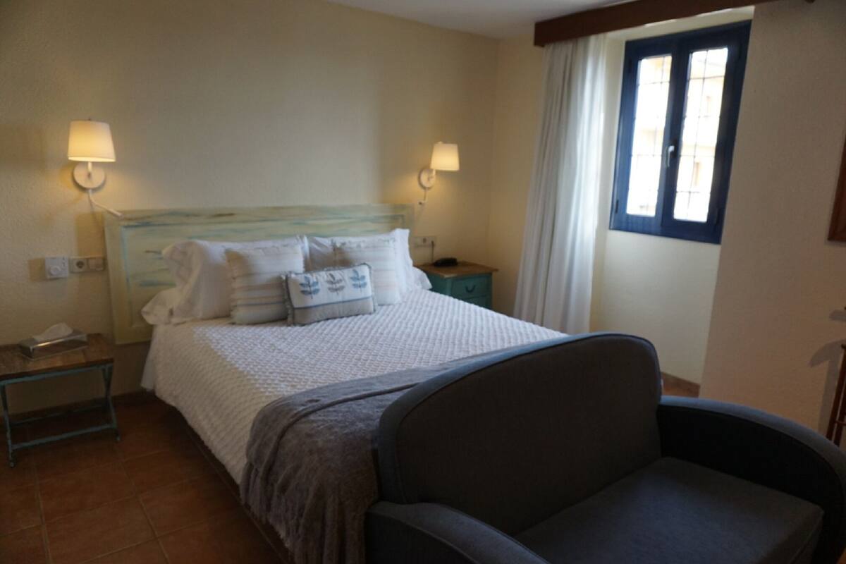 Une chambre d’hôtel avec un lit, deux lampes de chevet, une table de chevet, une chaise et une fenêtre avec des rideaux.