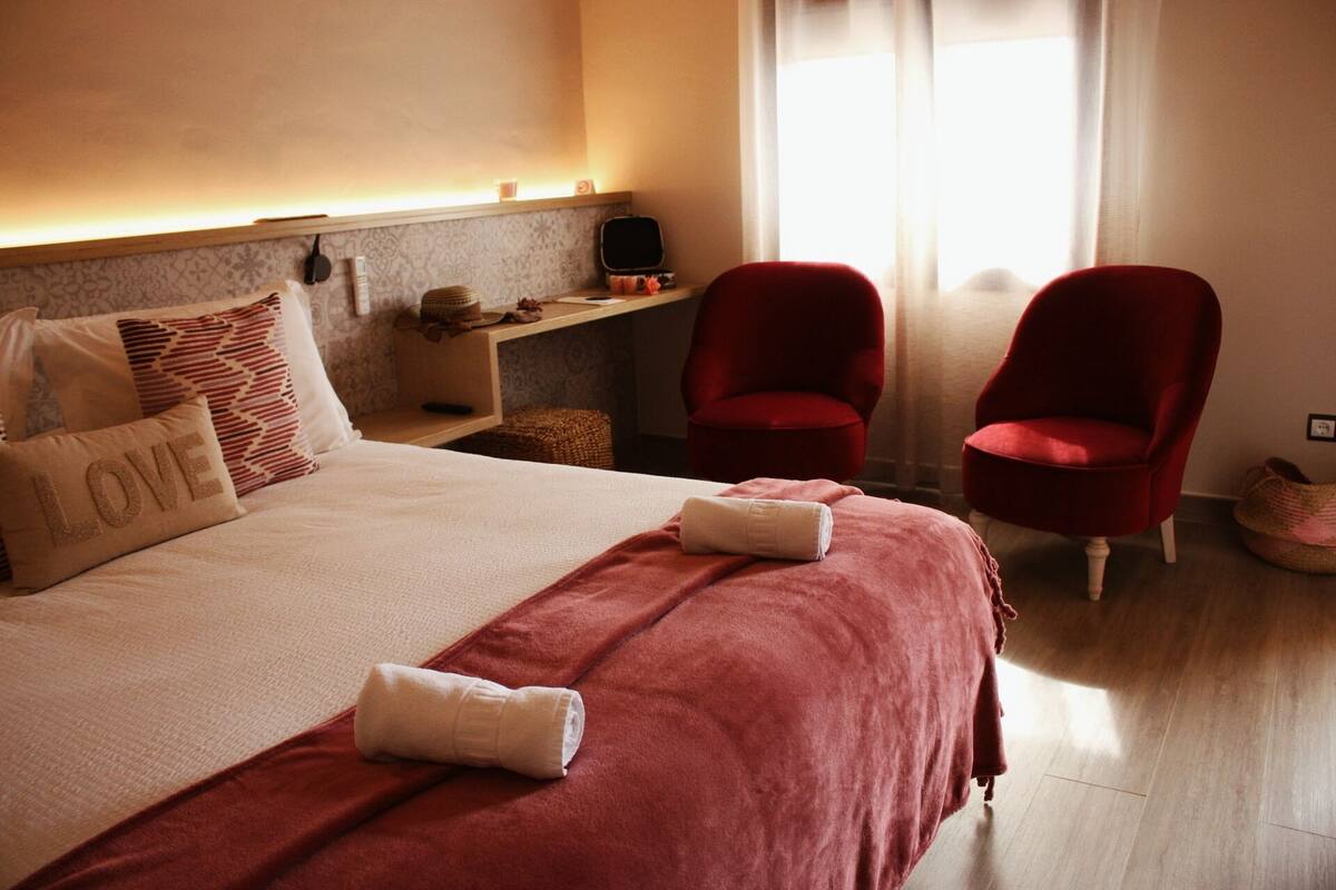 Une chambre d’hôtel avec un lit, deux fauteuils rouges, une petite table avec une lampe et une fenêtre avec des rideaux.