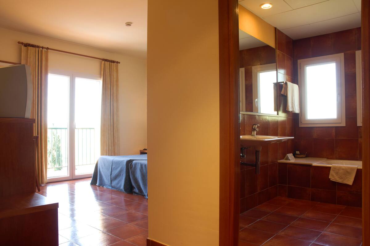 Une chambre d’hôtel avec un balcon, une salle de bain avec un lavabo et un miroir, et un lit recouvert d’une couverture bleue.