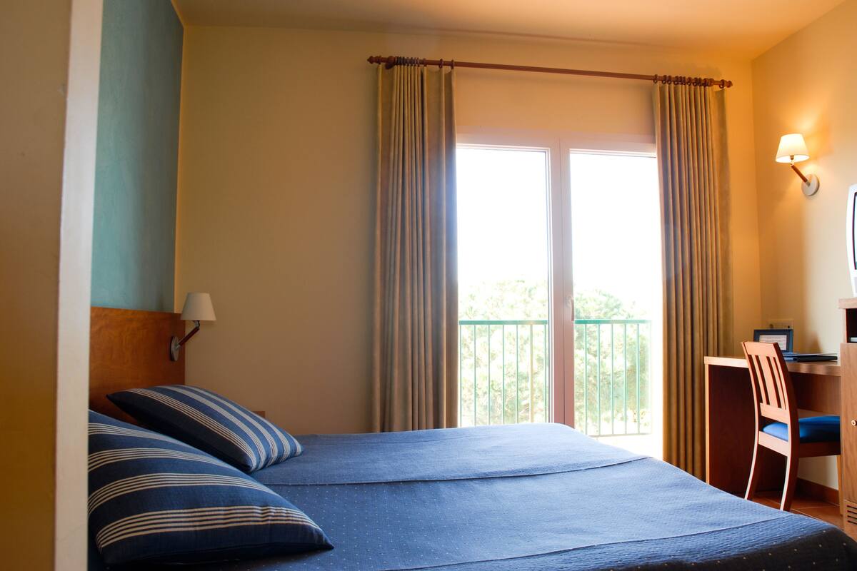Une chambre d’hôtel avec un lit, un bureau, une chaise et une fenêtre avec des rideaux.