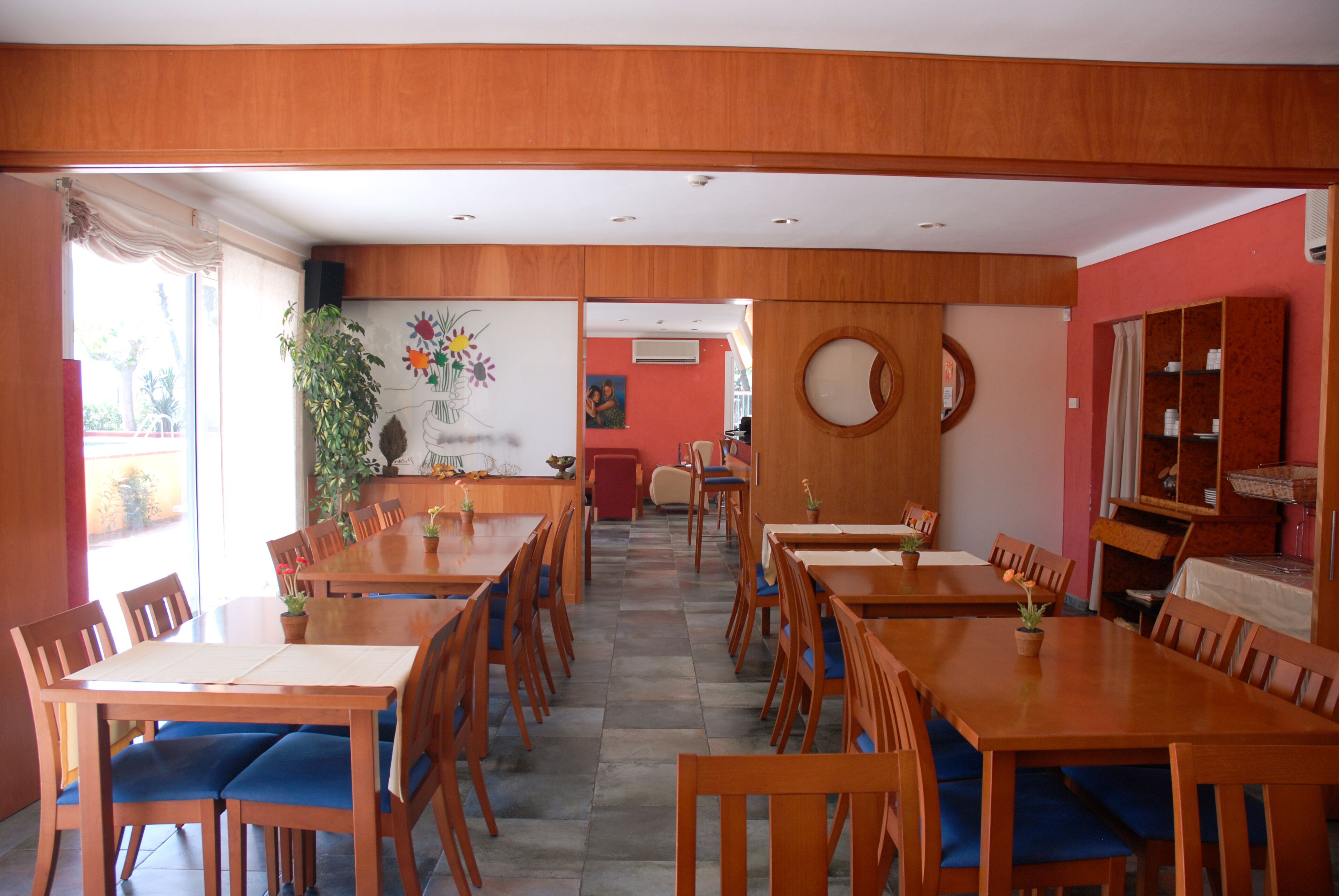 Un comedor con mesas y sillas de madera, plantas en macetas y vistas al exterior.