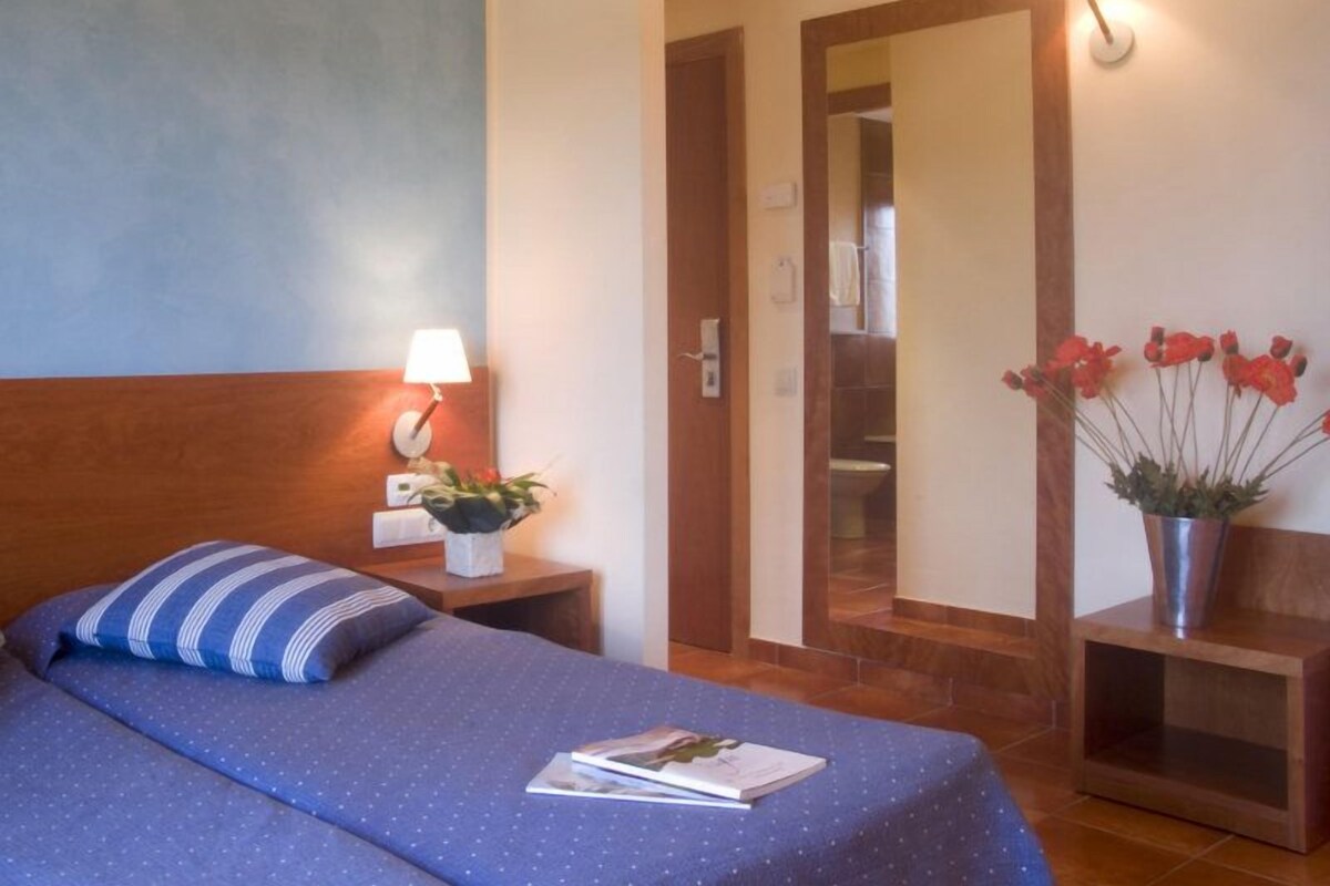 Ein Hotelzimmer mit einem Bett, einem Nachttisch mit Lampe, einem Spiegel und einer Vase mit Blumen.