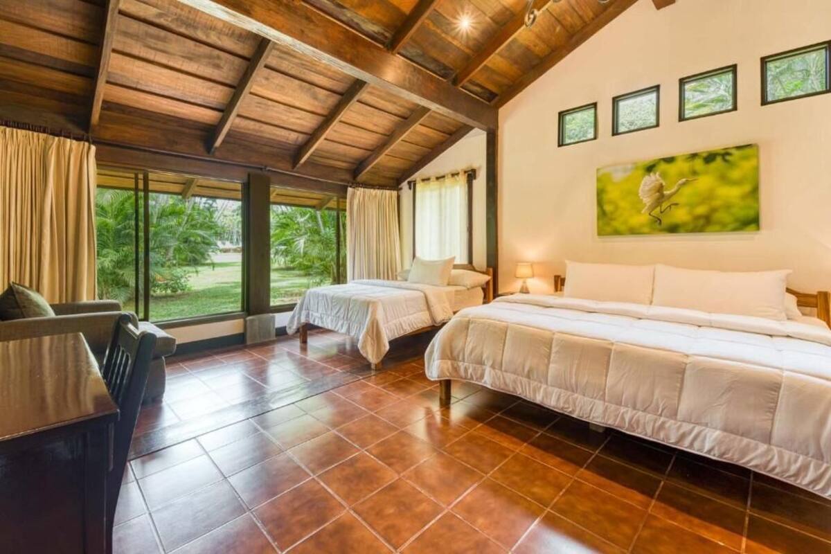 A room with two beds, a wooden ceiling, large windows, and a painting on the wall.