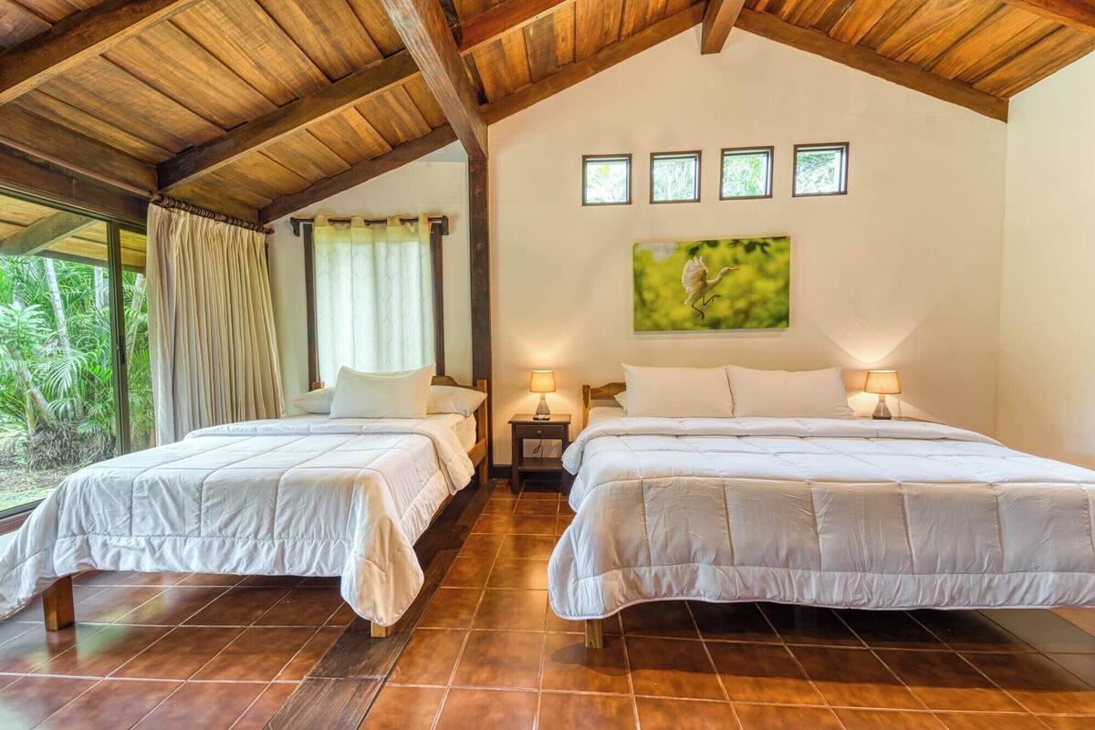 A room with two beds, wooden flooring, a sloped ceiling, and a large window with curtains.