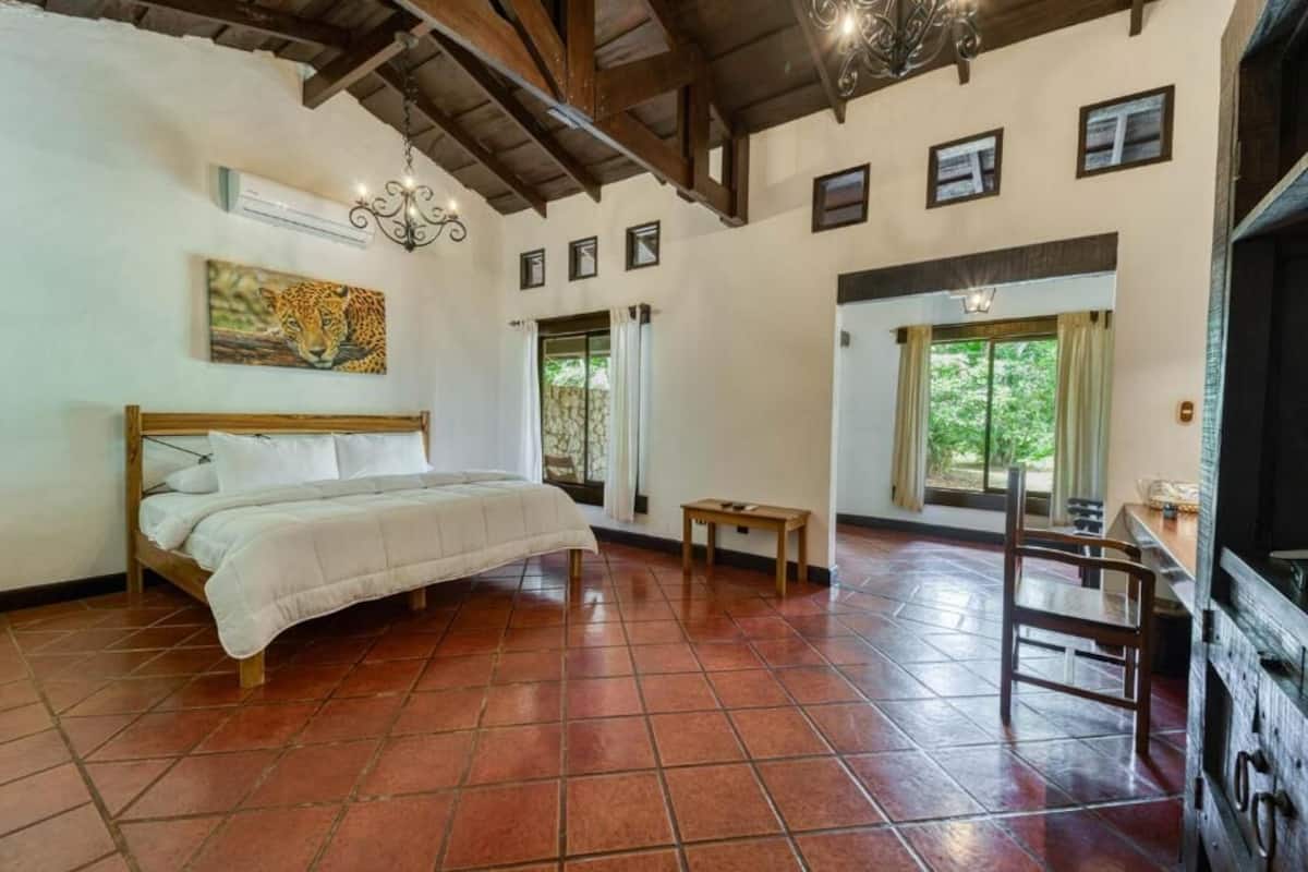 A bedroom with a bed, a small table, a chair, a painting, and a window.