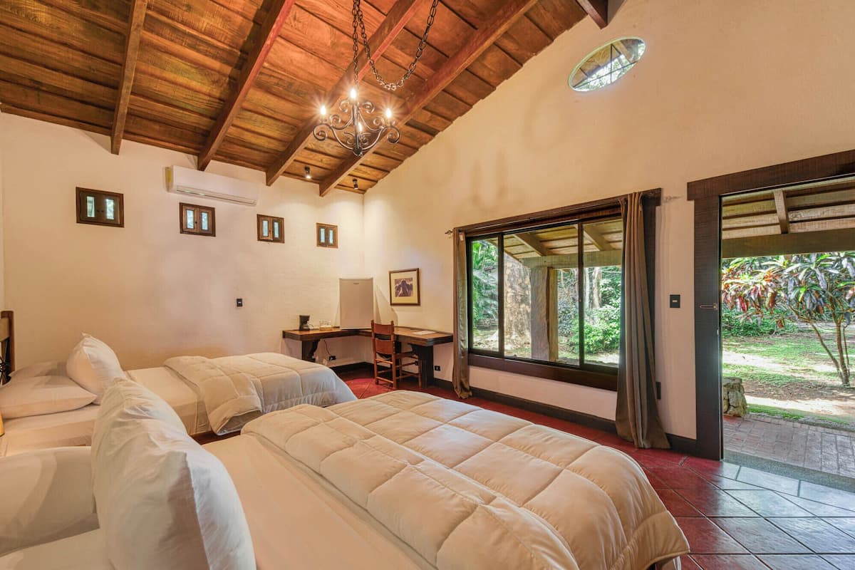 A bedroom with two beds, a desk, a chair, a chandelier, and a large window with a view of greenery.