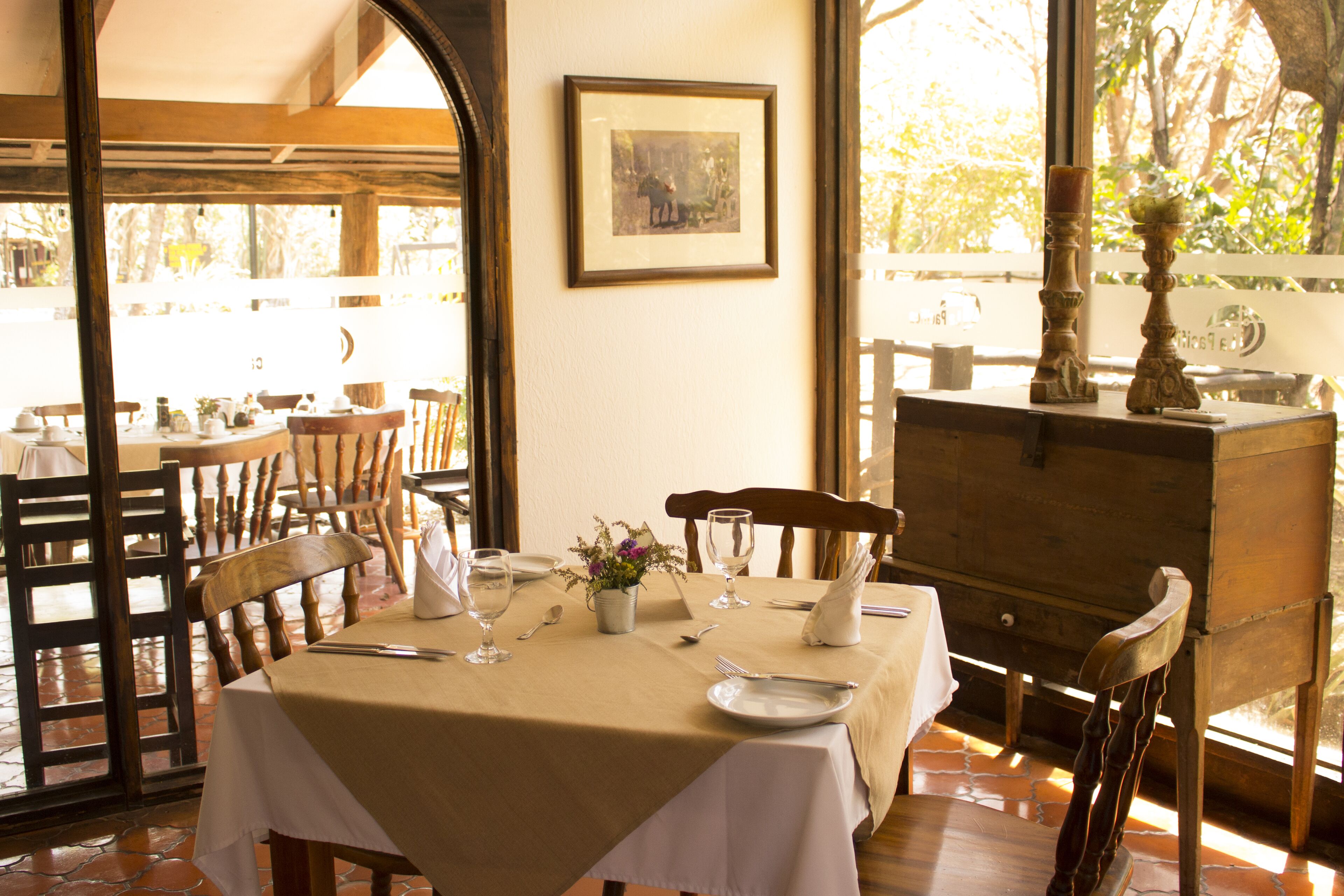 Un comedor con una mesa dispuesta para dos, sillas de madera, un cuadro en la pared y vistas al exterior.