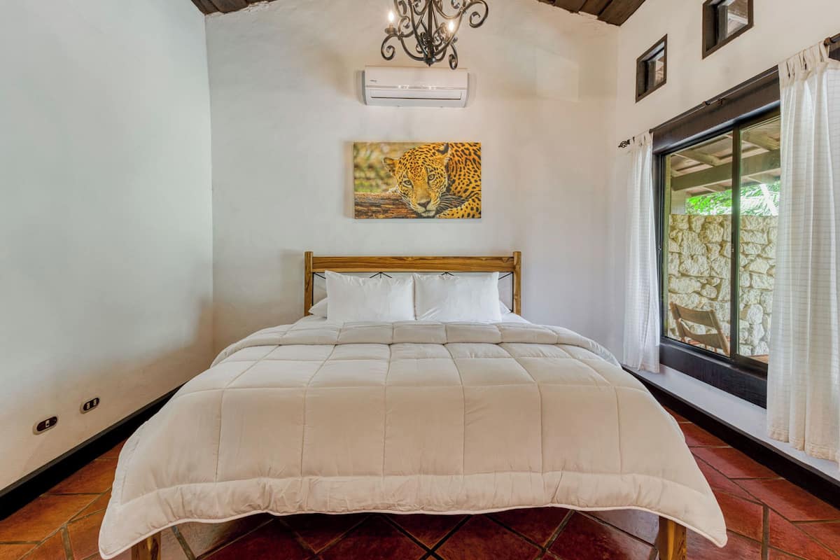 A bedroom with a wooden bed, white bedding, a leopard painting on the wall, and a stone wall visible through the window.