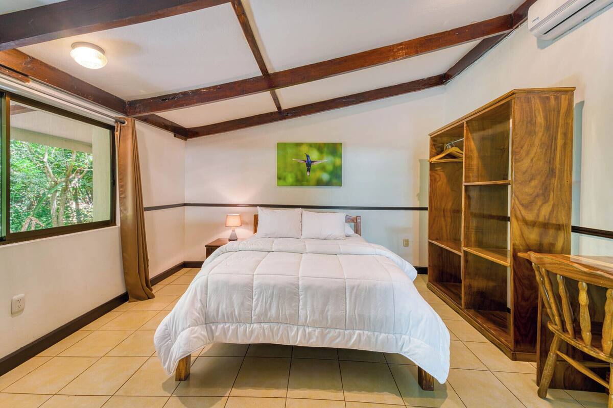 A bedroom with a large bed, a nightstand, a wooden wardrobe, and a window with a view of trees.