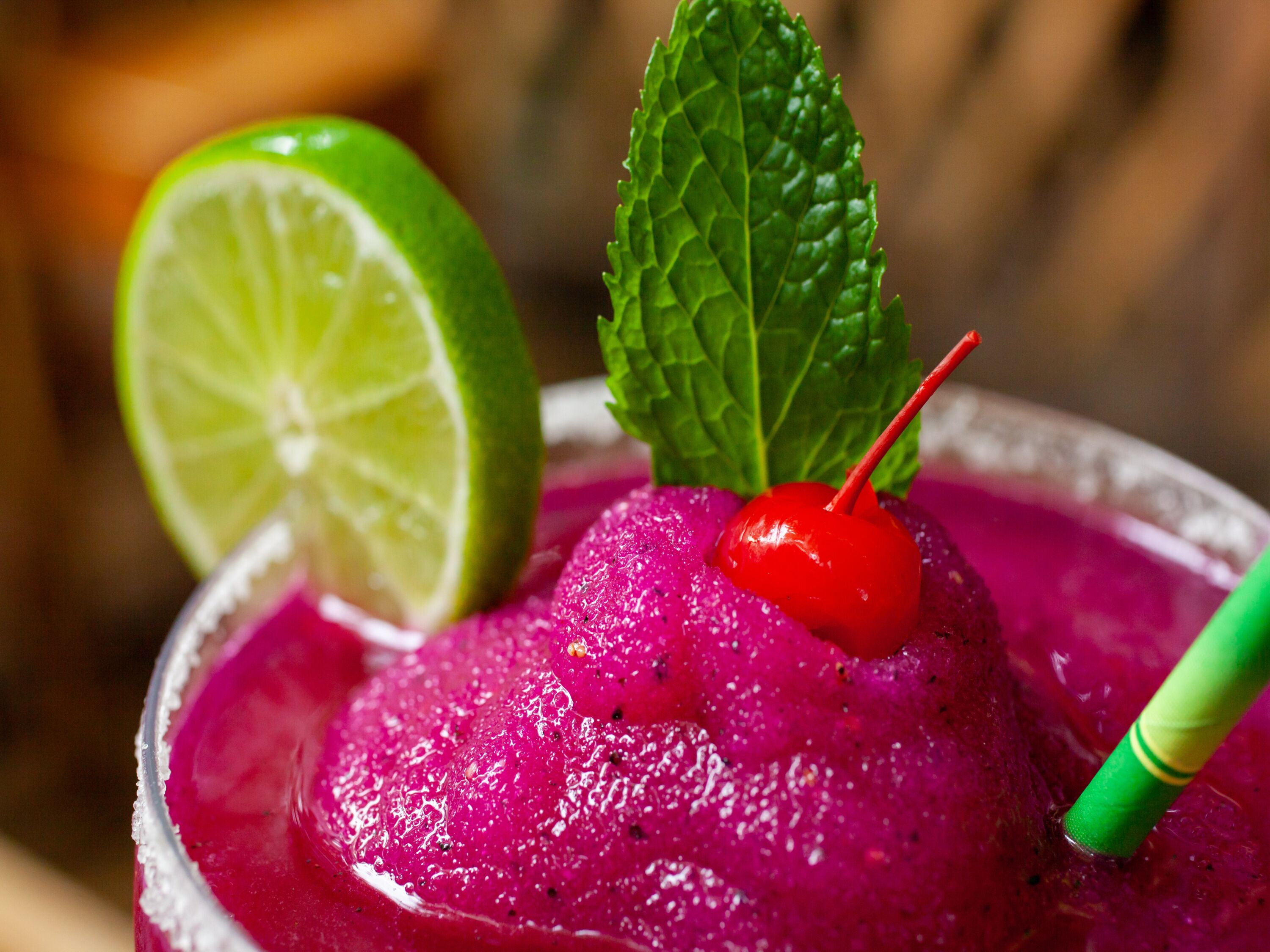 A close-up of a vibrant purple drink garnished with a lime wedge, a cherry, and a mint leaf.
