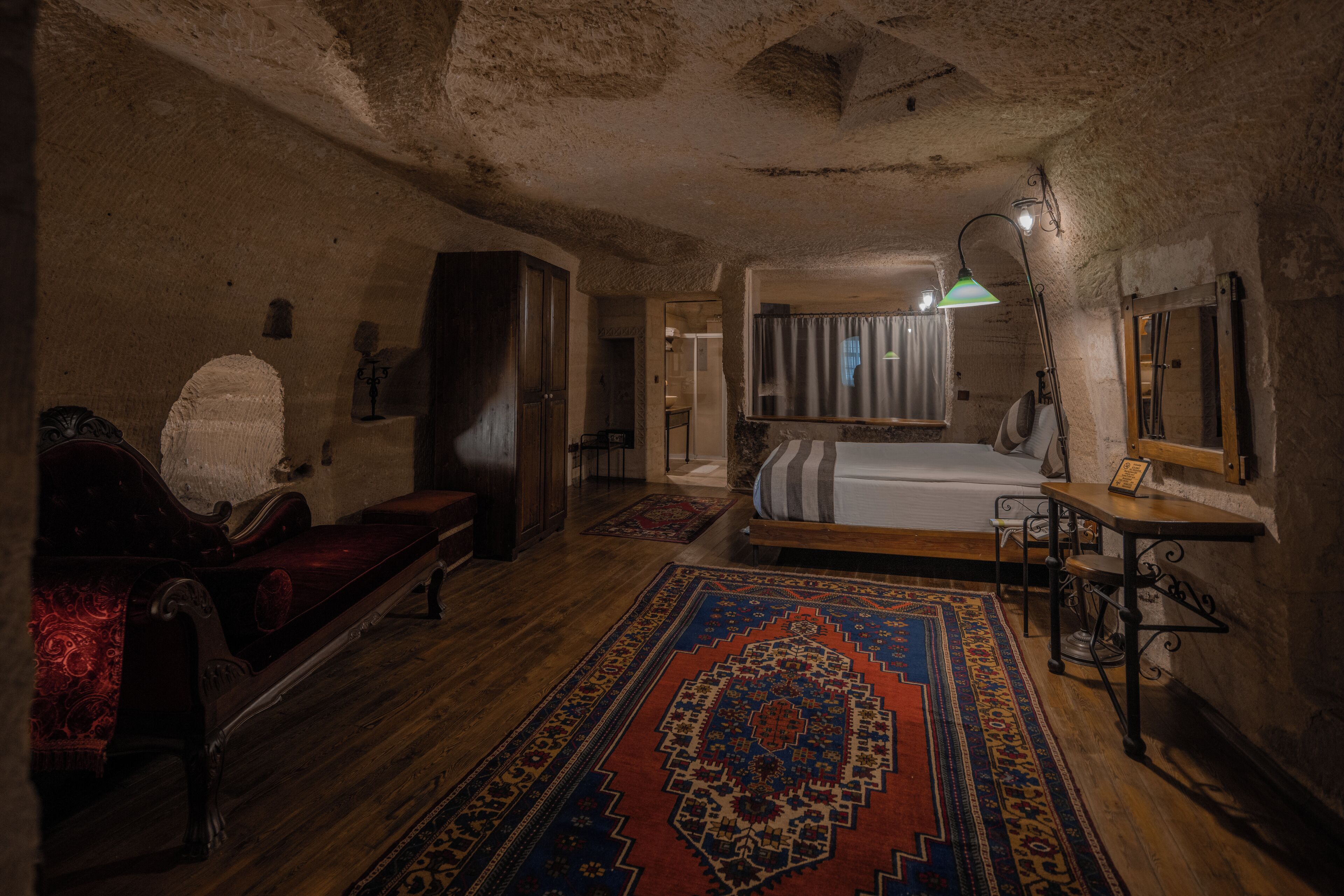 A room with a wooden floor, a large bed, a desk, a chair, a bench, and a rug.