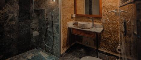 Une salle de bain avec une douche en pierre, un miroir encadré en bois, un lavabo avec un plan de toilette en marbre et des toilettes.