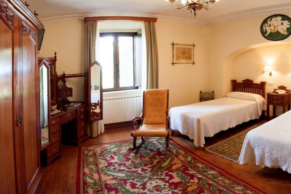 A traditional bedroom with two single beds, a wooden wardrobe, a chair, and a large window with curtains.
