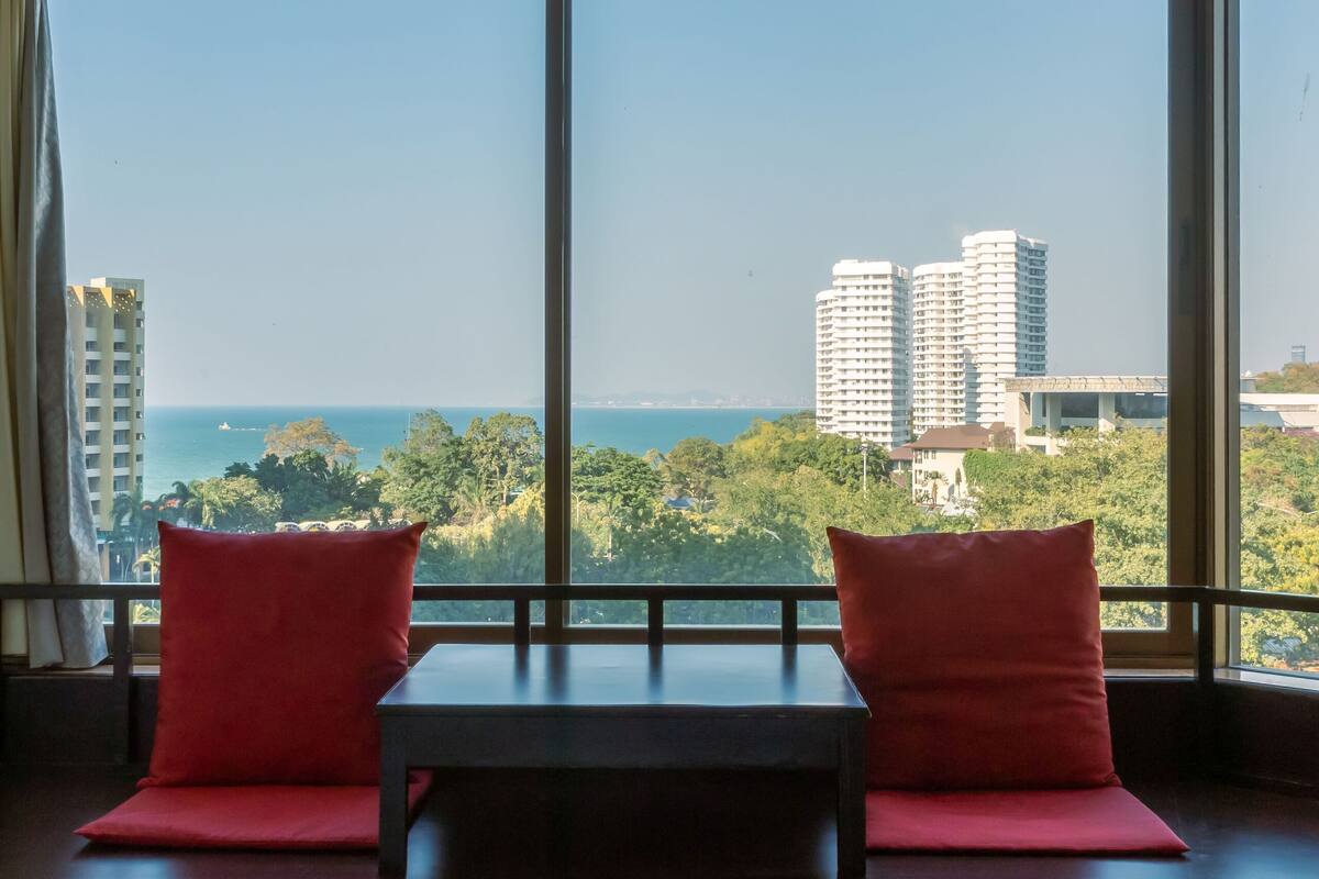 A room with a large window, two red pillows, a small table, and a view of the city and ocean.