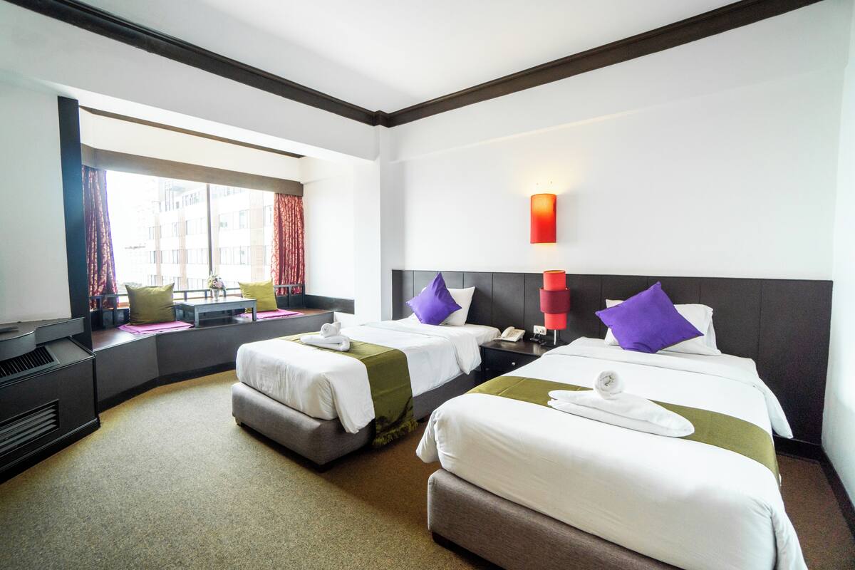 A hotel room with two beds, a desk, a chair, a window with curtains, and a wall-mounted lamp.