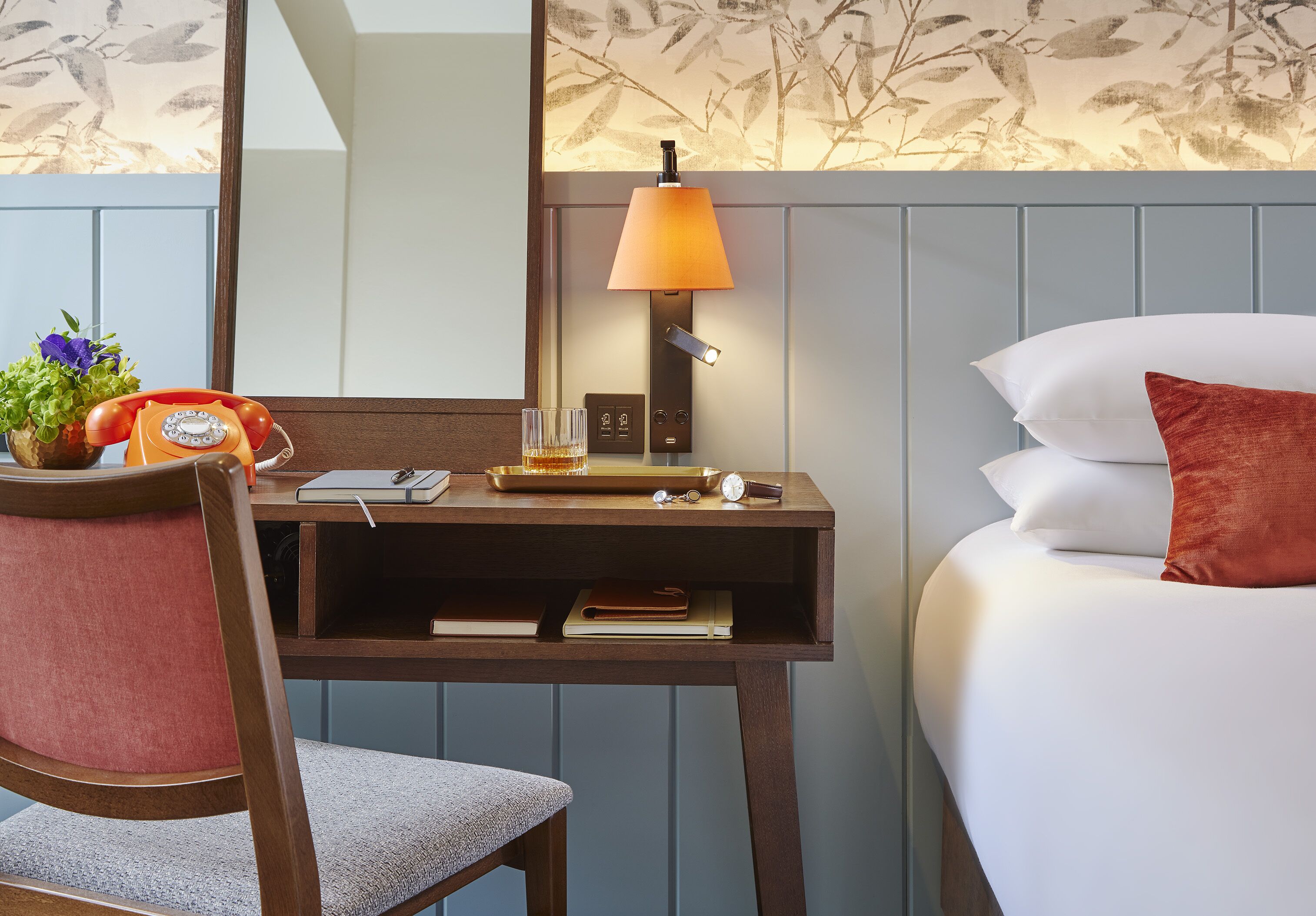 A hotel room with a wooden desk, a telephone, a lamp, and a bed with white and red pillows.