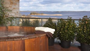 A wooden hot tub with towels, surrounded by potted plants, overlooking a lake with a stone wall and a view of the distant shoreline. - Hodson Bay Hotel (Athlone)