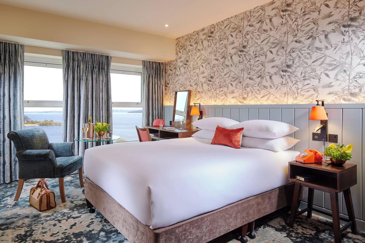 A hotel room with a large bed, a desk with a chair, a small table with a lamp, and a view of the sea.