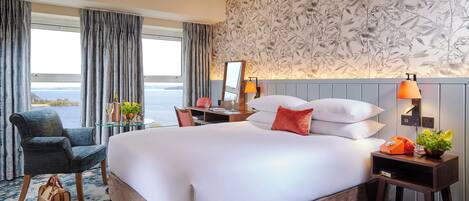A hotel room with a large bed, a desk with a chair, a small table with a lamp, and a view of the sea.