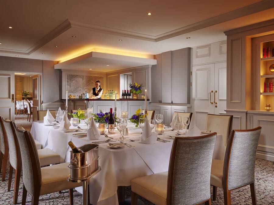 Une salle à manger élégante, avec une longue table dressée pour un repas, un coin bar avec un barman et une bibliothèque agrémentée d’objets décoratifs.