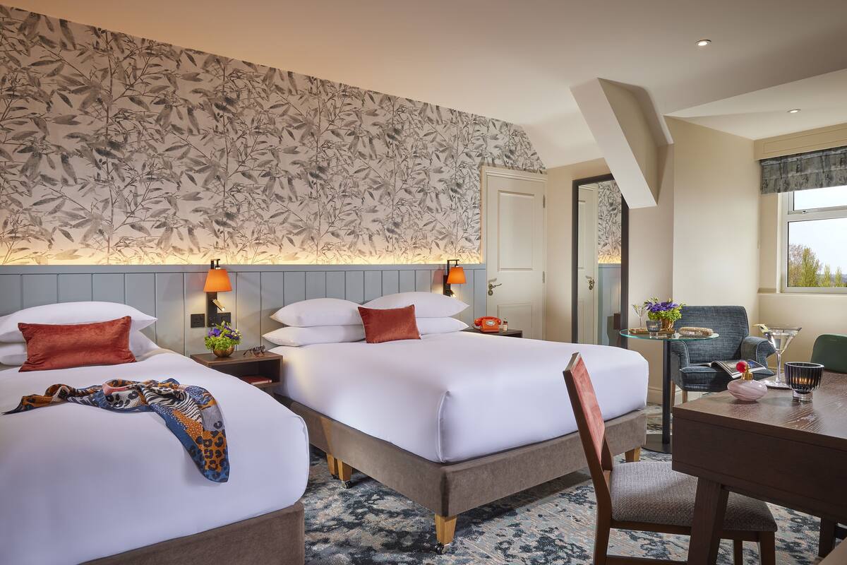 A hotel room with two beds, a dining table, and a chair. There is a patterned wallpaper on the wall, a nightstand with a lamp, and a small table with a vase of flowers.