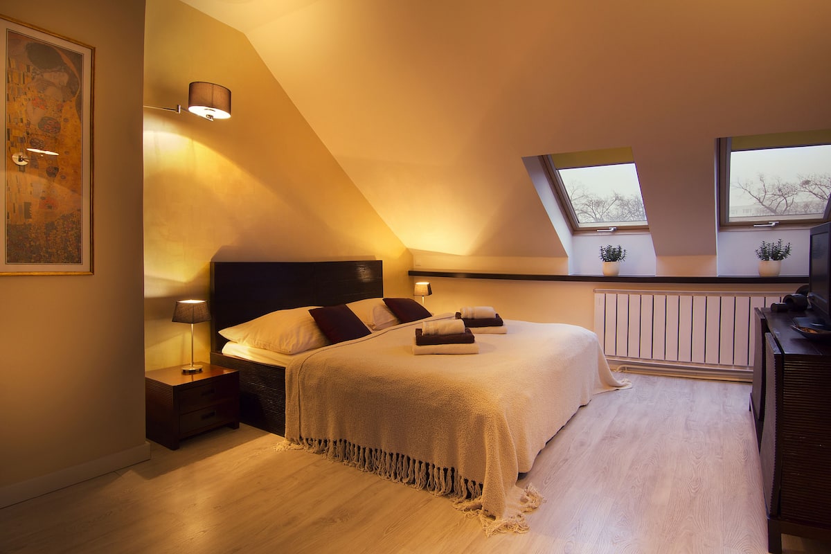 A bedroom with a sloped ceiling, a bed with a white bedspread, two bedside lamps, a nightstand, a dresser, a radiator, and two windows with plants.