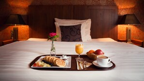 Un lit bien fait, avec un plateau de petit-déjeuner comprenant des viennoiseries, des fruits et un verre de jus d’orange, dans une chambre d’hôtel dotée d’une tête de lit à motifs et de lampes de chaque côté.