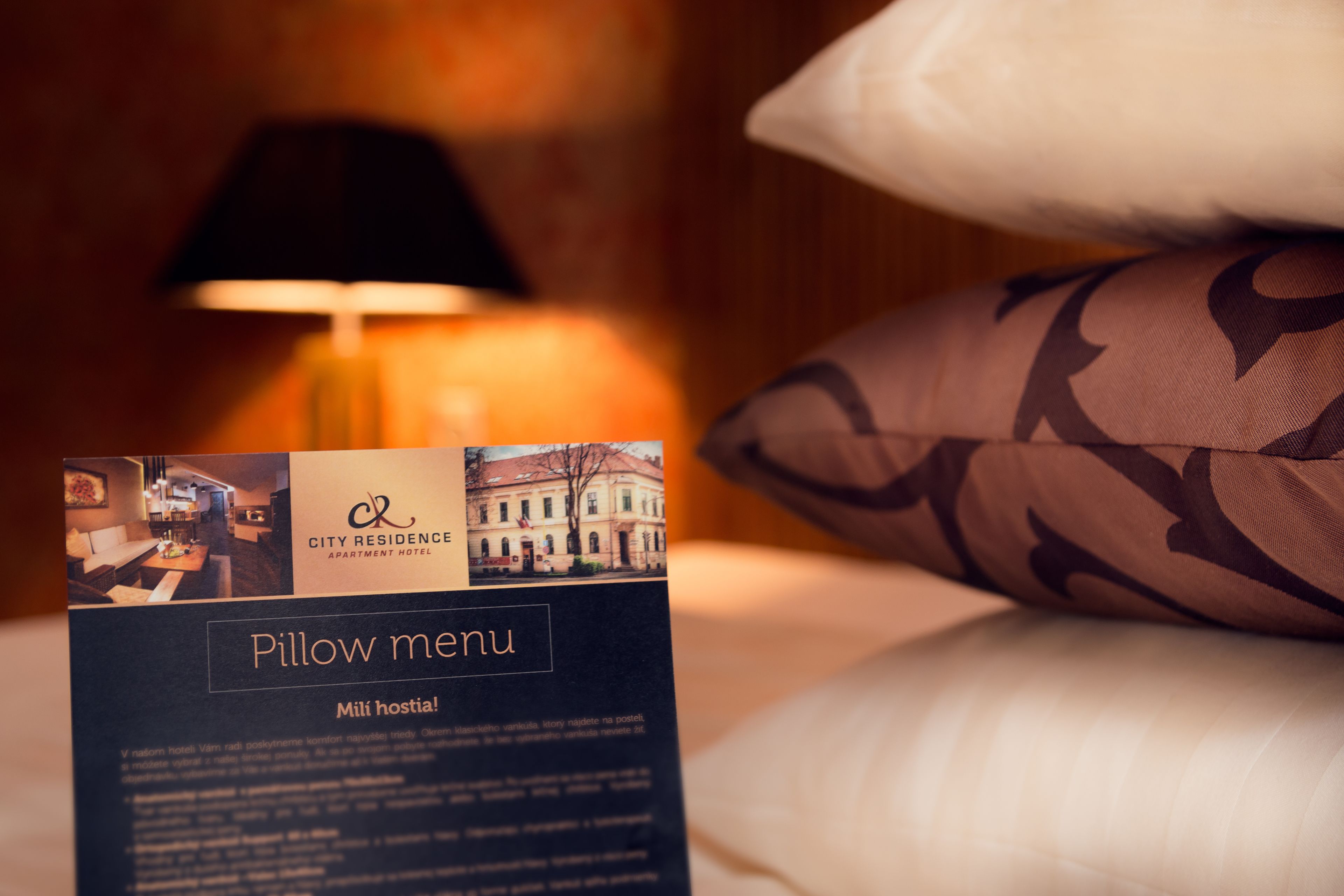 Carta de almohadas del Apart Hotel City Residence, mostrando una cama con almohadas estampadas y una lámpara al fondo.