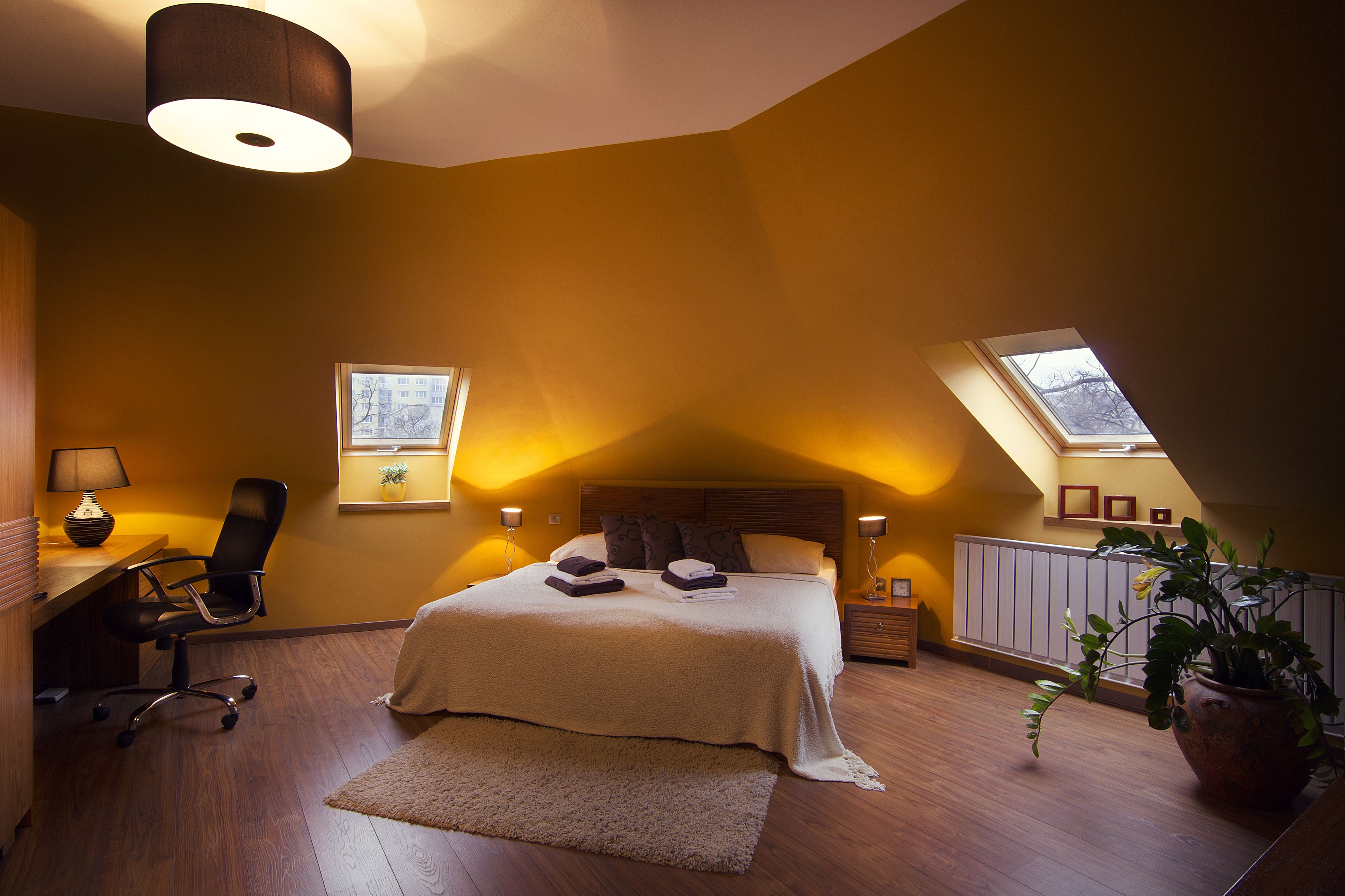 A bedroom with a large bed, a desk with a chair, a potted plant, and a skylight.