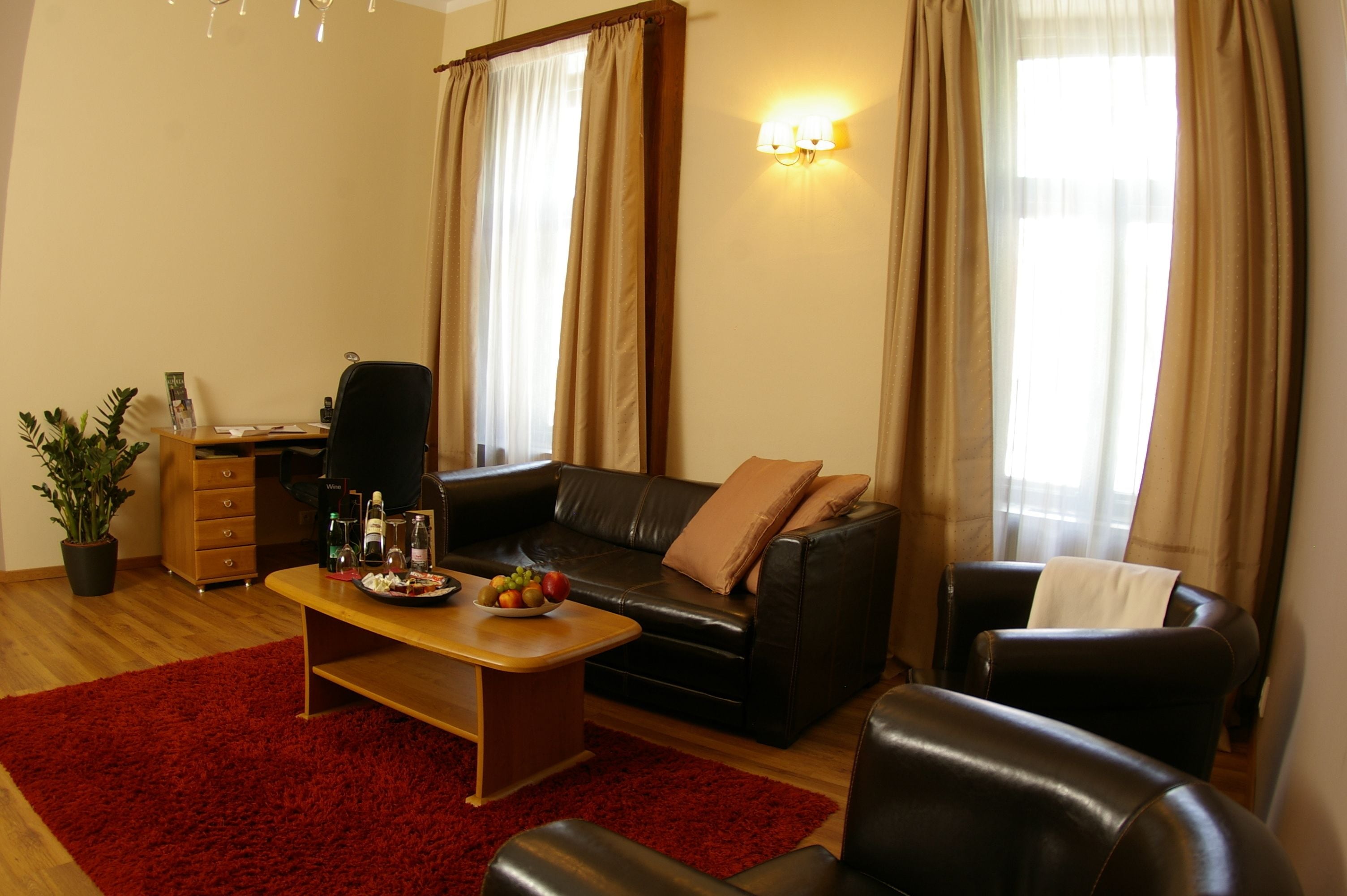 A living room with a black leather sofa, a wooden coffee table, and a red rug. There is a desk with a chair and a potted plant.