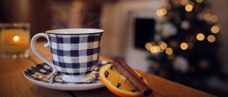 A checkered cup on a saucer with a slice of orange and a cinnamon stick, steam rising from the cup, set on a wooden table with a blurred background featuring a lit candle and a Christmas tree.