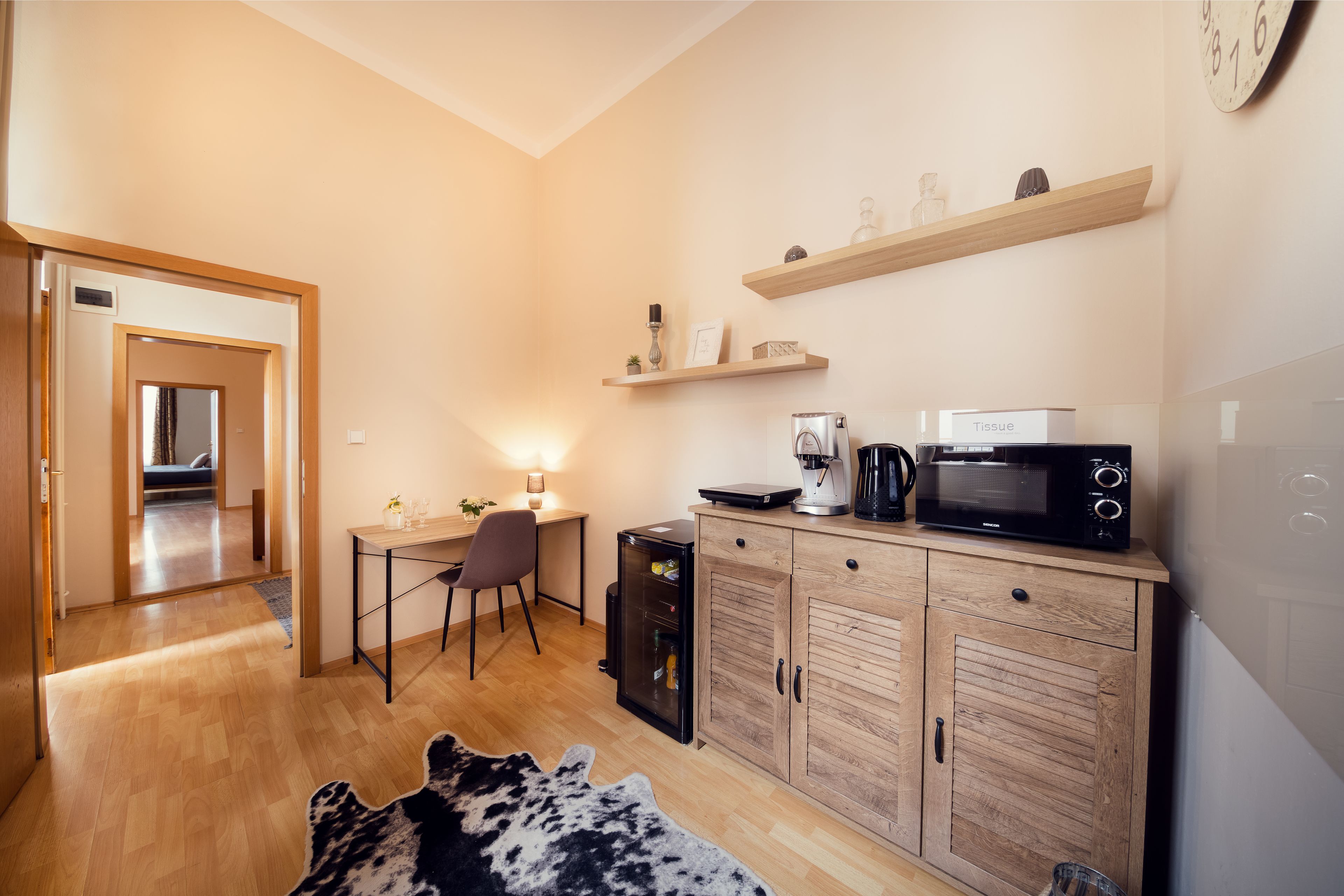 A compact kitchen with a microwave, coffee maker, and a small refrigerator. There is a wooden cabinet with a shelf above it, and a desk with a chair. A cowhide rug is on the floor.