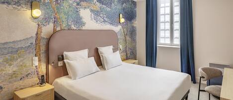 A hotel room with a large bed, a nightstand with a lamp, a desk with a chair, and a window with curtains.