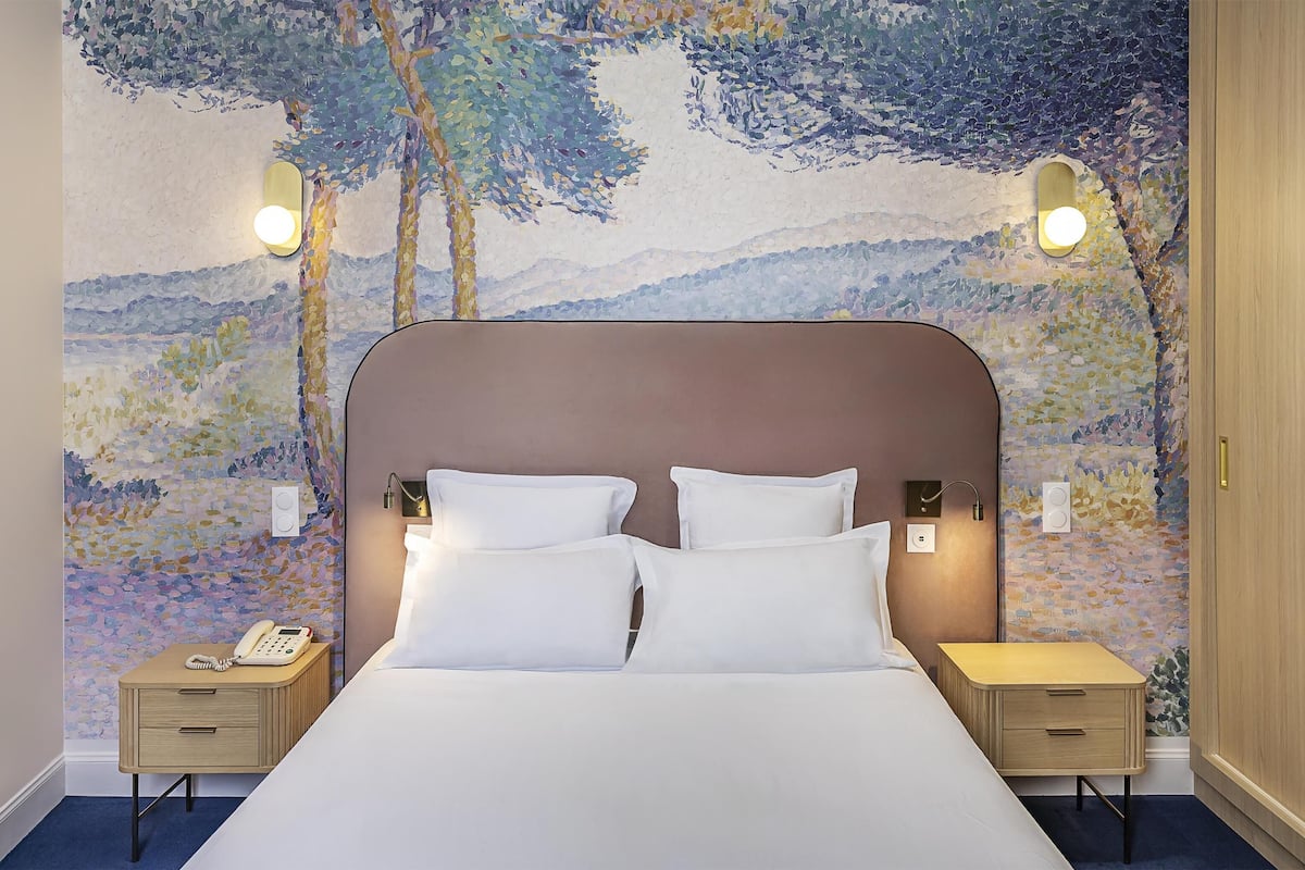 Una habitación de hotel con una cama grande, dos mesitas de noche y una pared con un paisaje pintado.