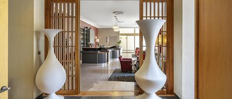 A modern hotel lobby with a wooden reception desk, two large white vases, and a view into the dining area.