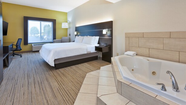 A hotel room with a bed, a desk, a chair, a large window, a wall-mounted TV, and a bathtub.