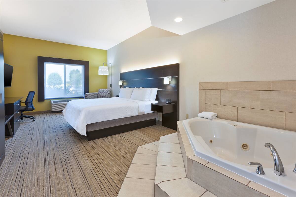 A hotel room with a bed, a desk, a chair, a large window, a wall-mounted TV, and a bathtub.