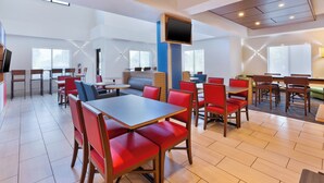 A modern hotel dining area with multiple tables and chairs, a flat-screen TV mounted on a blue pillar, and large windows providing natural light.