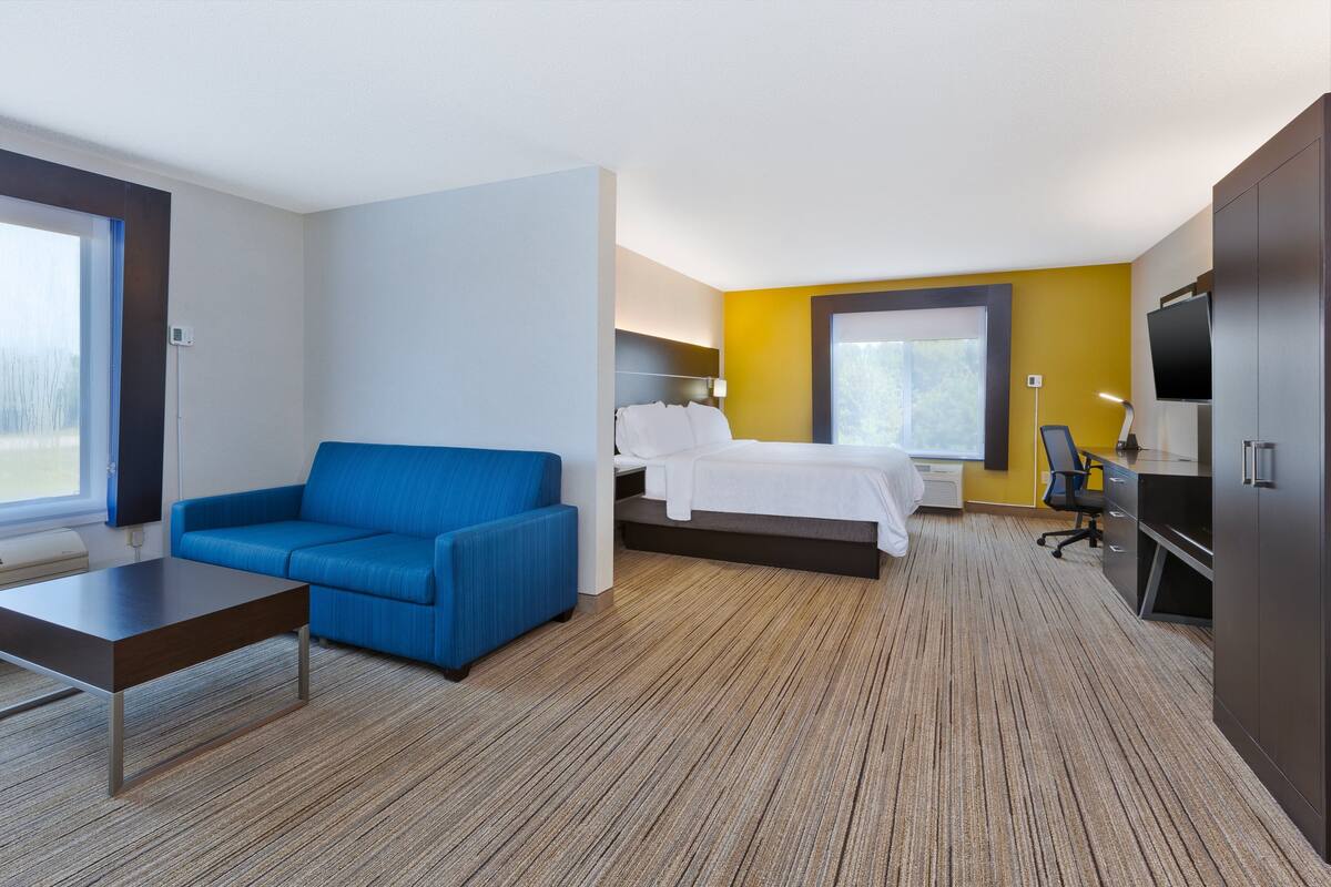A modern hotel room with a blue sofa, a bed with white bedding, a desk with a chair, and a TV.
