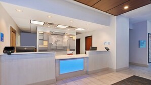 A hotel reception area with a modern design, including a reception desk, digital check-in station, and a sign indicating guest room locations.