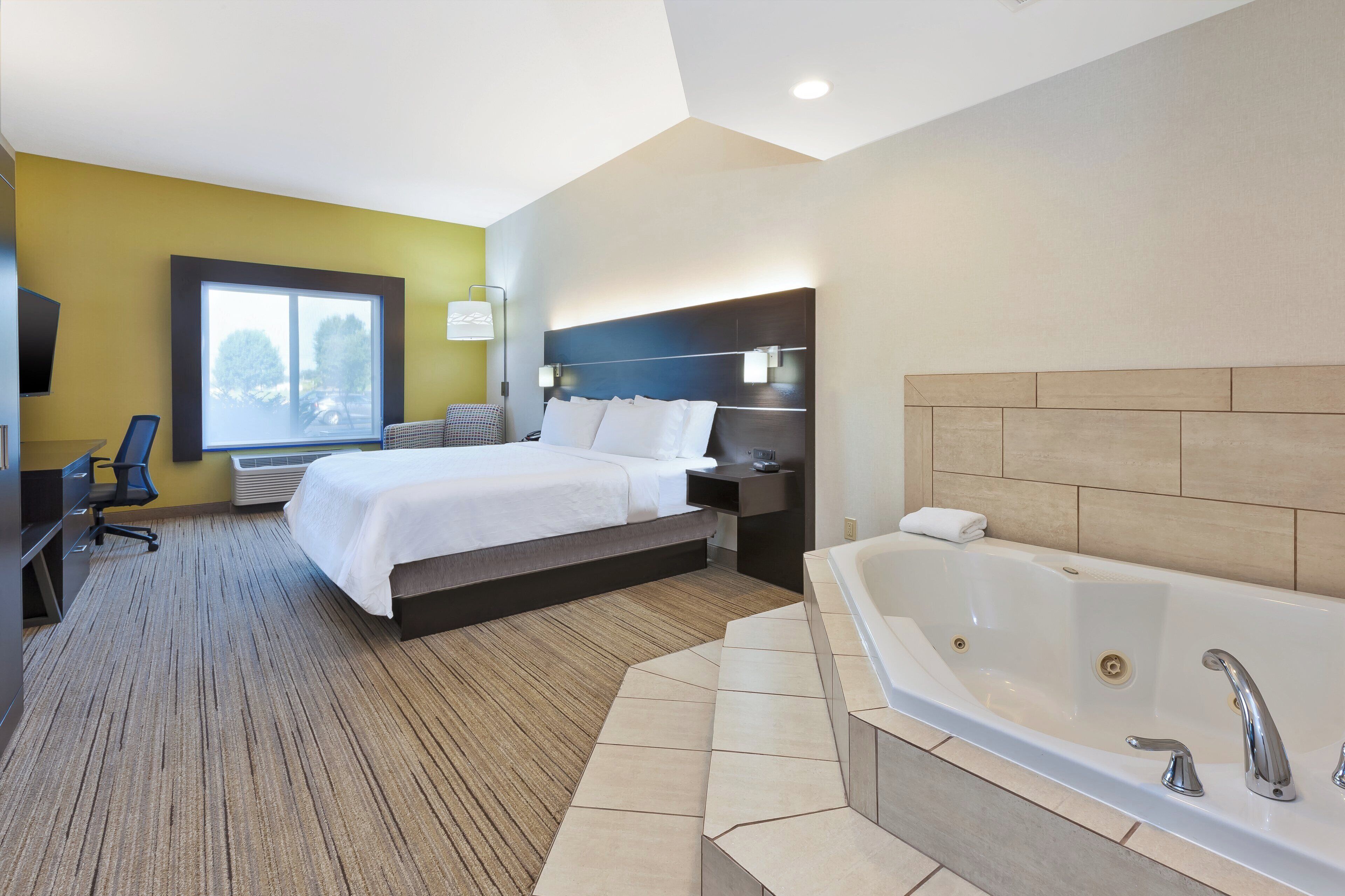 A hotel room with a bed, a desk, a chair, a large window, a wall-mounted TV, and a bathtub.